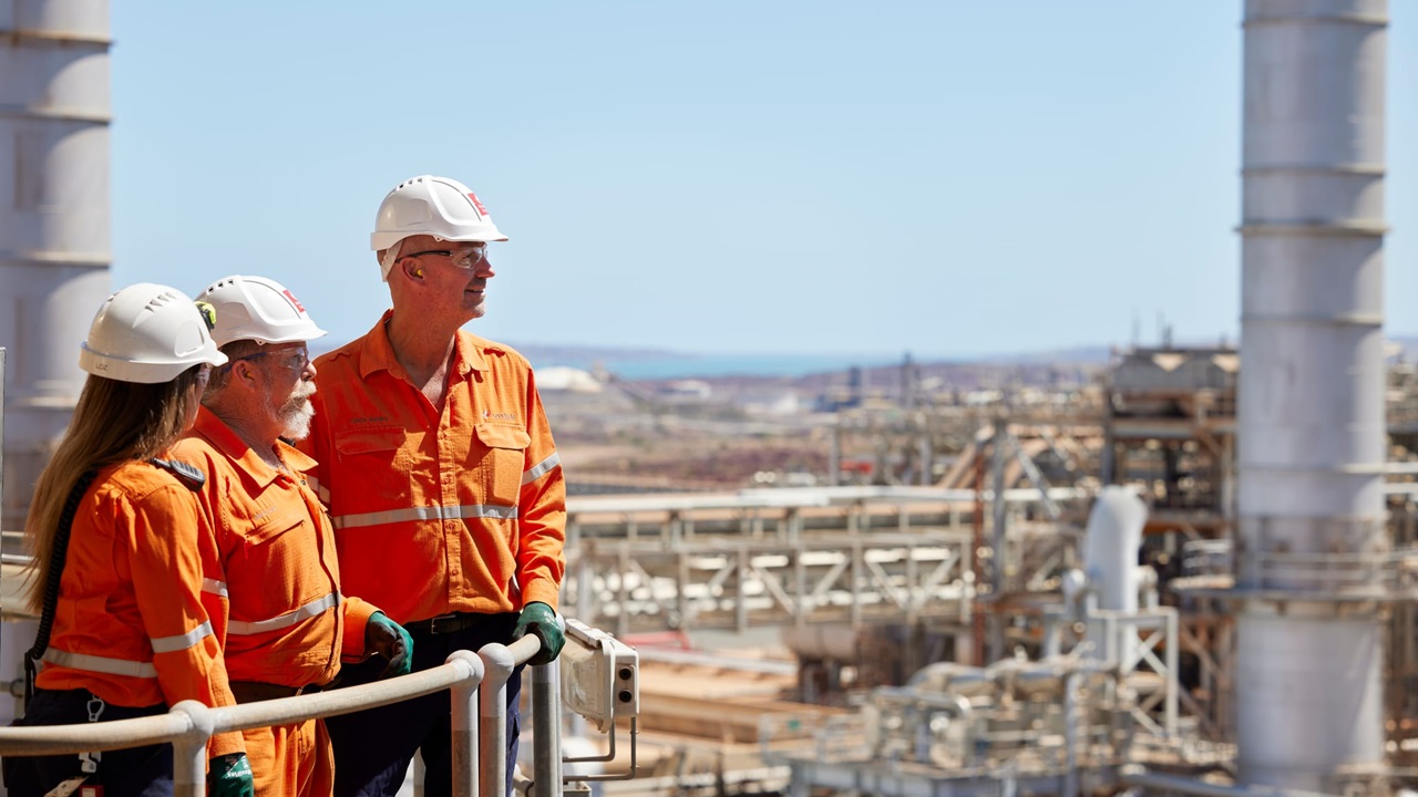 Three Woodside employees in high-vis and hard hats, standing on a platform looking out onto the Scarborough Gas Plant